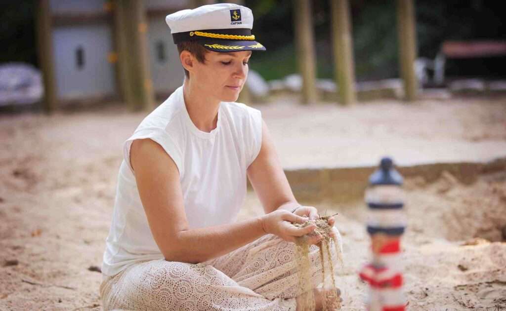Katharina Koll sitzt in einer Sandkiste mit Kapitänsmütze, lässt Sand sanft durch ihre Finger rieseln. Neben ihr steht ein kleiner Leuchtturm – Symbol für Orientierung, Ruhe und inneren Frieden.